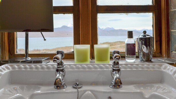A bathroom with a sink, two faucets, and a window with a view of a beach and mountains. - 1 bedroom accommodation in Fearnmore, near Applecross (Fearnmore, near Applecross)