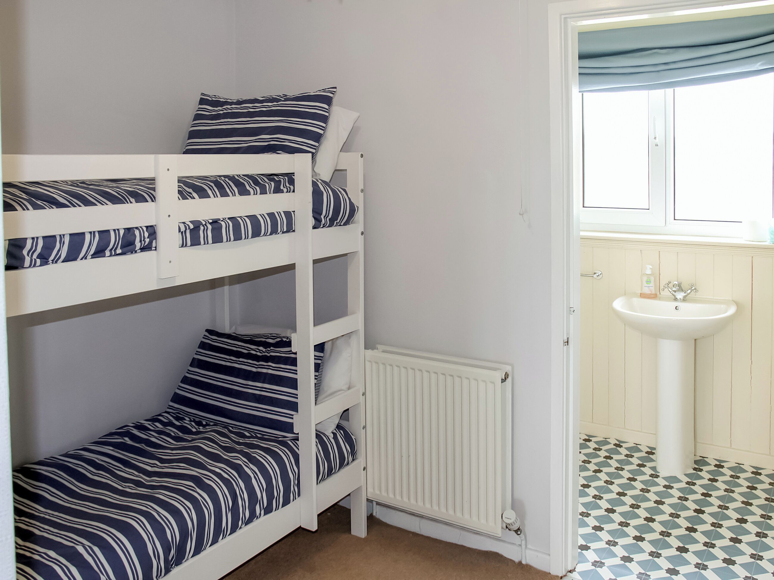 En køjeseng med stribet sengetøj, en hvid radiator, samt et badeværelse med vask og gulvfliser med mønster.