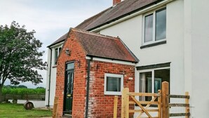 Une maison à deux étages avec une extension en briques, une porte en bois et une brouette dans la cour.