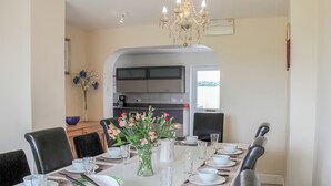 Un coin repas avec une longue table dressée pour le repas, des chaises, un vase avec des fleurs et, au fond, une cuisine.