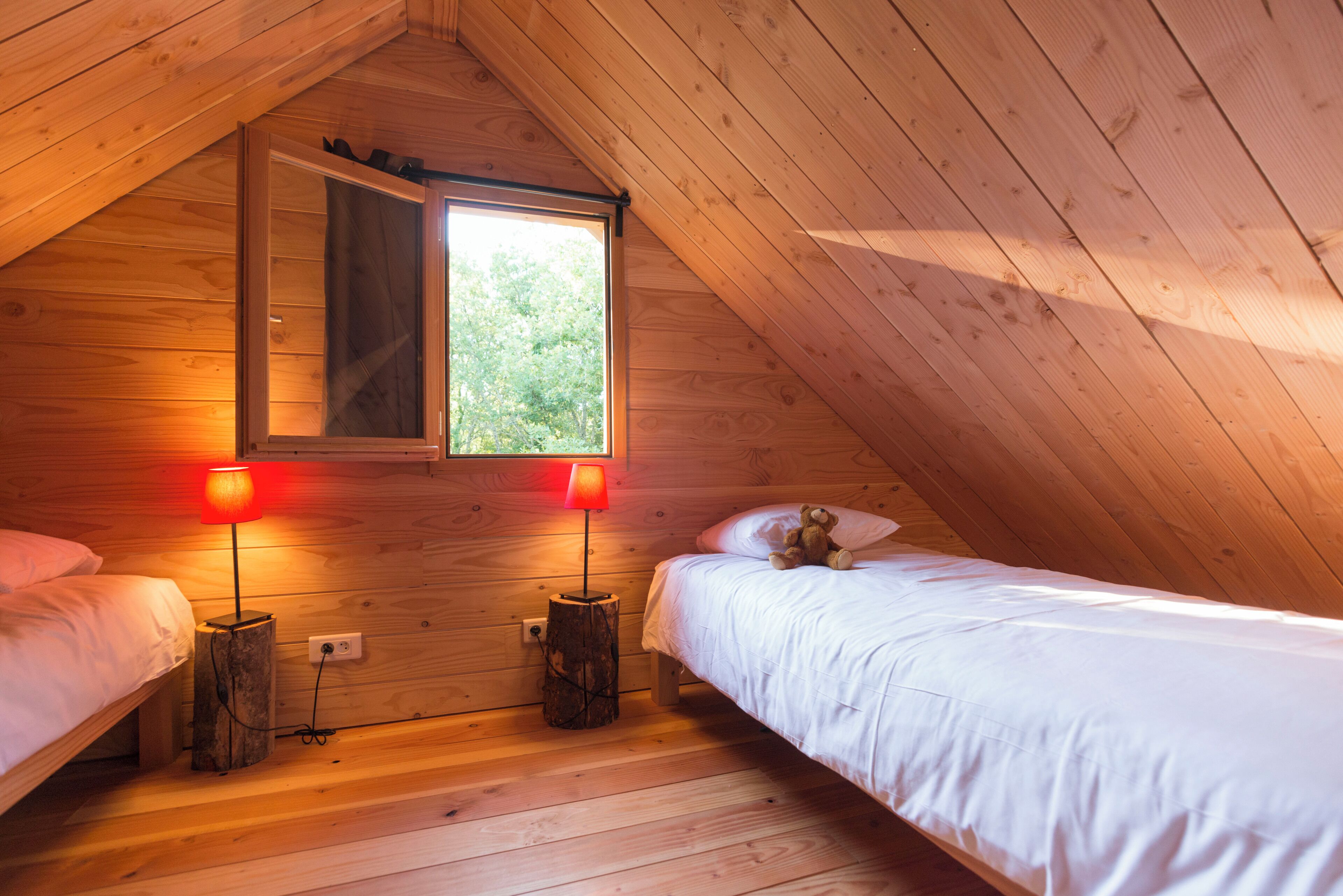 A cozy attic bedroom with wooden walls, a sloped ceiling, two beds, and a window letting in natural light.