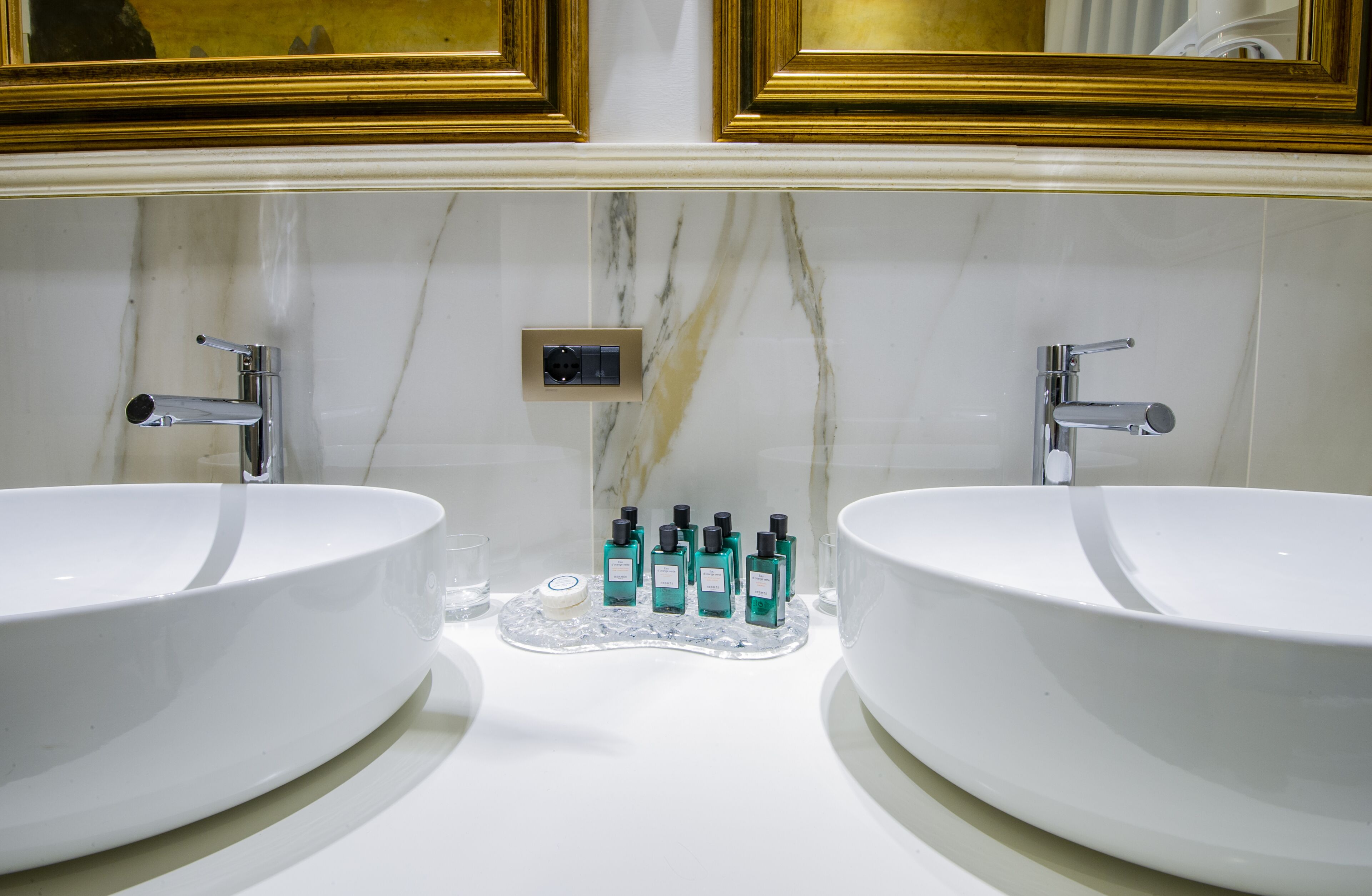 A bathroom with two large white sinks, a marble backsplash, and a gold-framed mirror above.