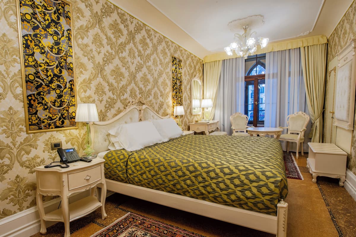 A classic bedroom with a large bed, bedside tables, a nightstand, a chair, a small table, and a mirror.