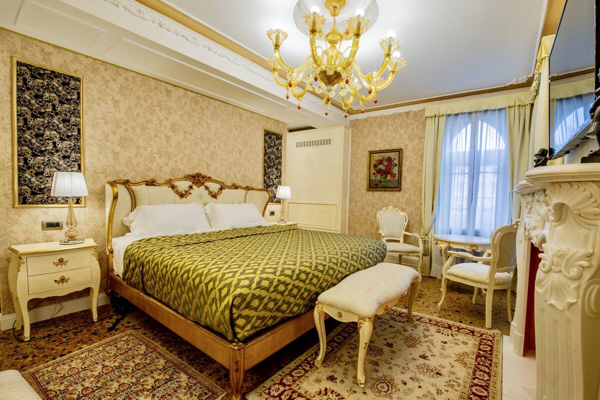 A classic bedroom with a large bed, a chandelier, a sitting area with a chair, and a decorative mirror.