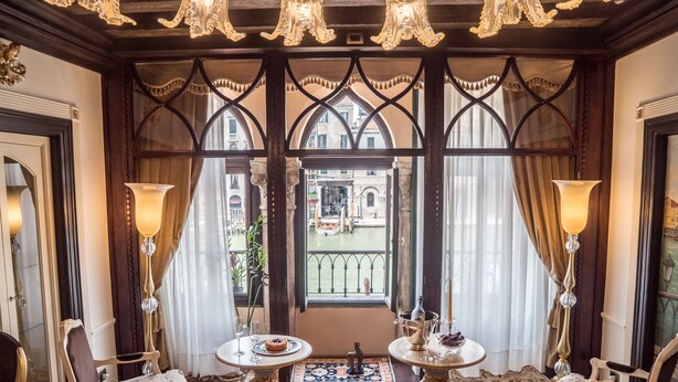 A room with a view of a canal, featuring a table with a cake, two chairs, and a decorative angel statue. - EGO’ Boutique Hotel - The Silk Road (Venice)