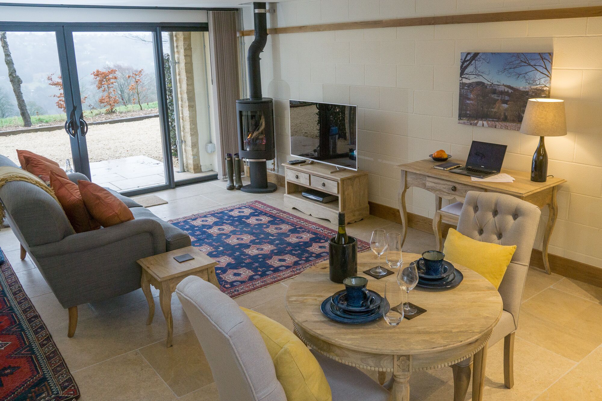 A living room with a sofa, a dining table with chairs, a television, a fireplace, and a laptop on a desk.