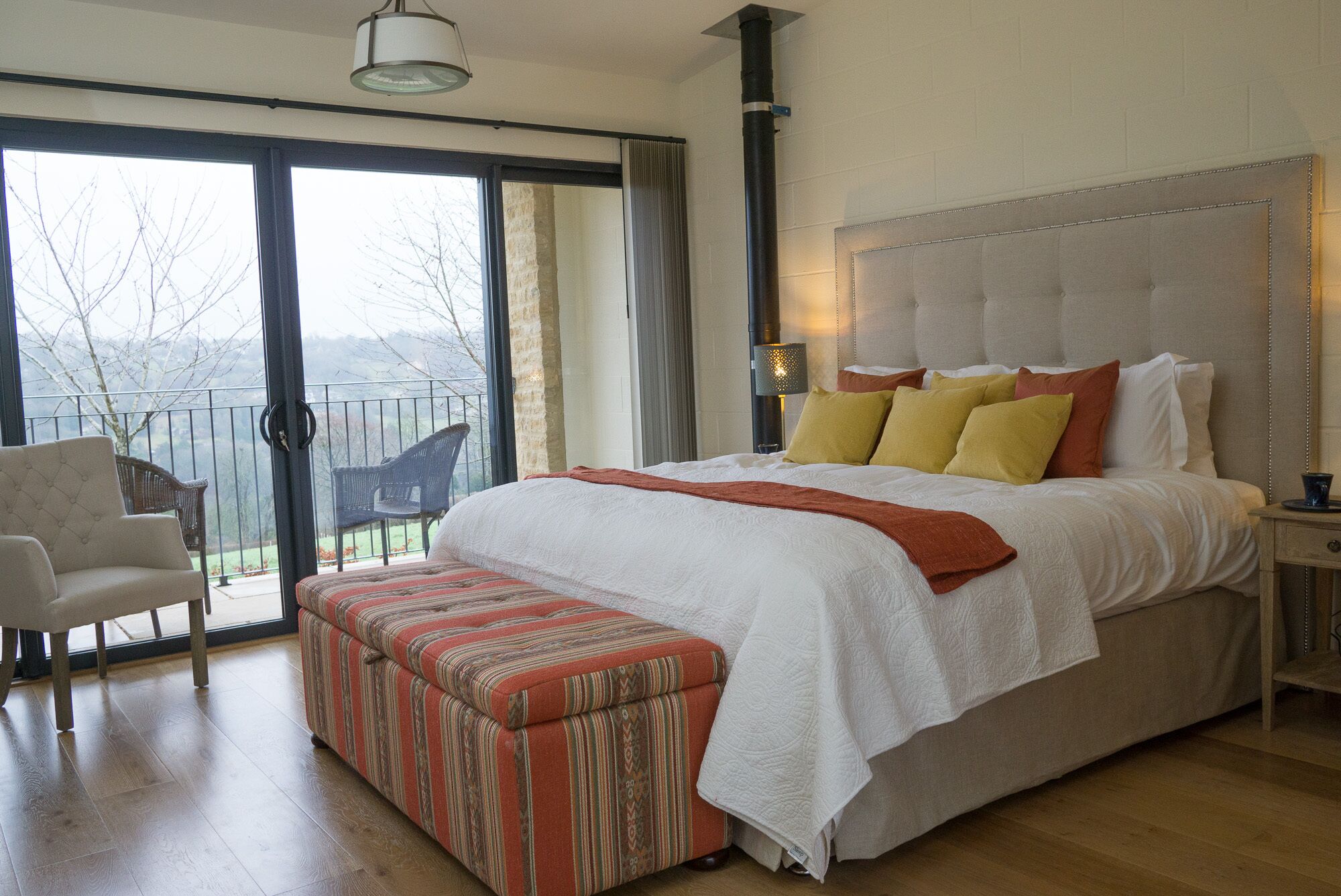 A large bed with a white comforter and orange throw, a wooden bench, a nightstand, and a chair.