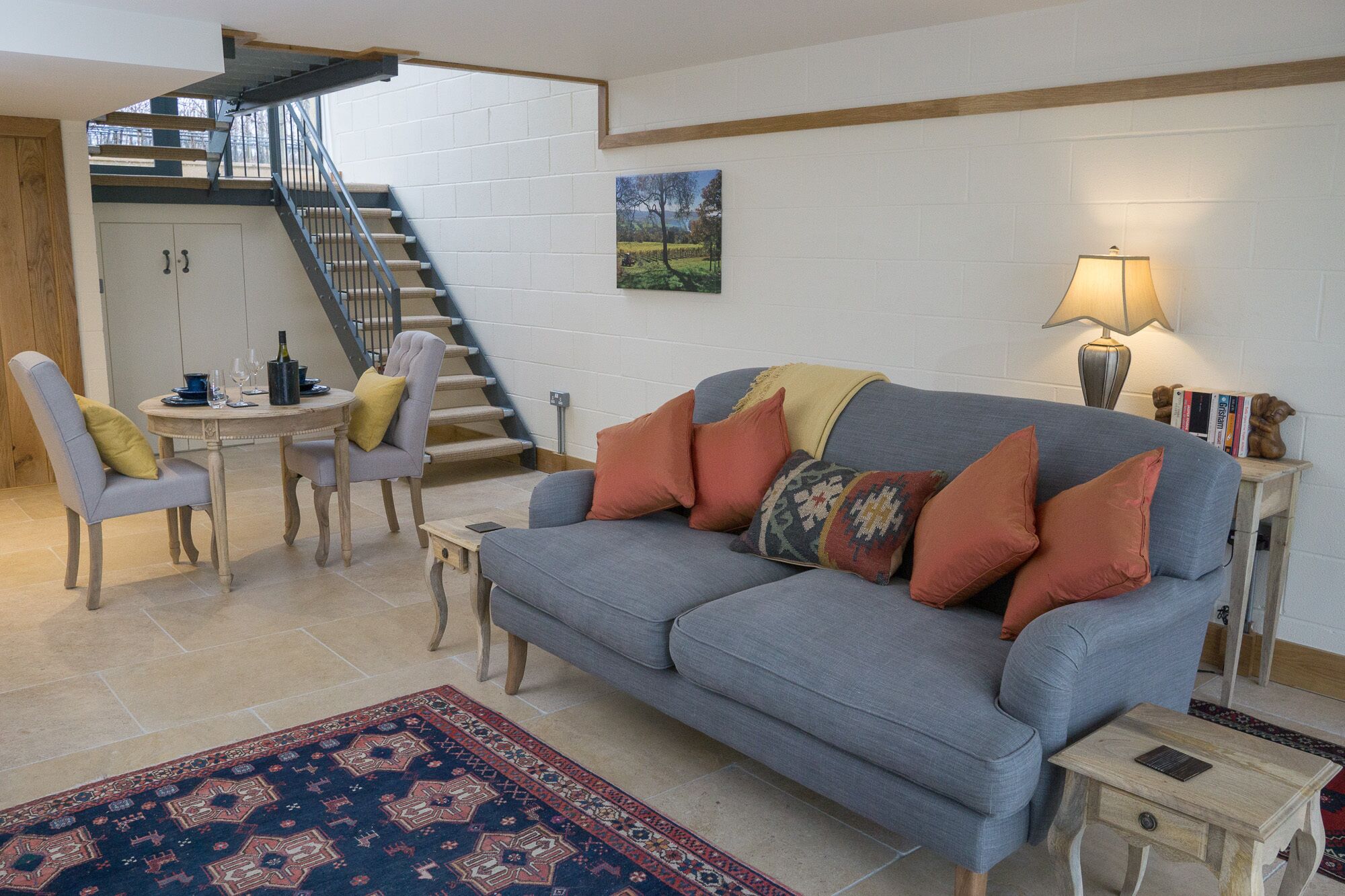 Un salon avec un canapé gris, un tapis à motifs, une table à manger avec des chaises, un escalier et un tableau accroché au mur.