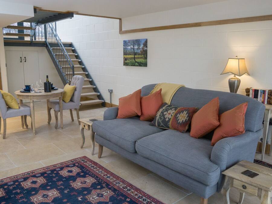 A living room with a grey sofa, a patterned rug, a dining table with chairs, a staircase, and a wall-mounted picture.