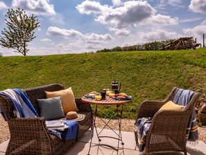 A wicker sofa with cushions and a table set with breakfast items, a hat, and a book, outdoors with a grassy hill and a wooden structure in the background. - Woodchester Valley Vineyard Barns (Stroud)