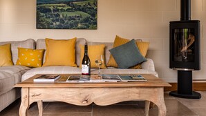Un salon avec un canapé, des coussins, une table basse avec des magazines, une bouteille de vin et une cheminée.