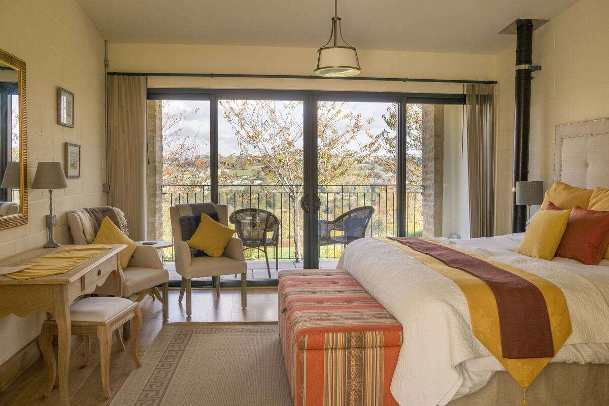 Ett hotellrum med en säng, ett skrivbord, en stol, en soffa och en balkong med utsikt över träd och ett staket.