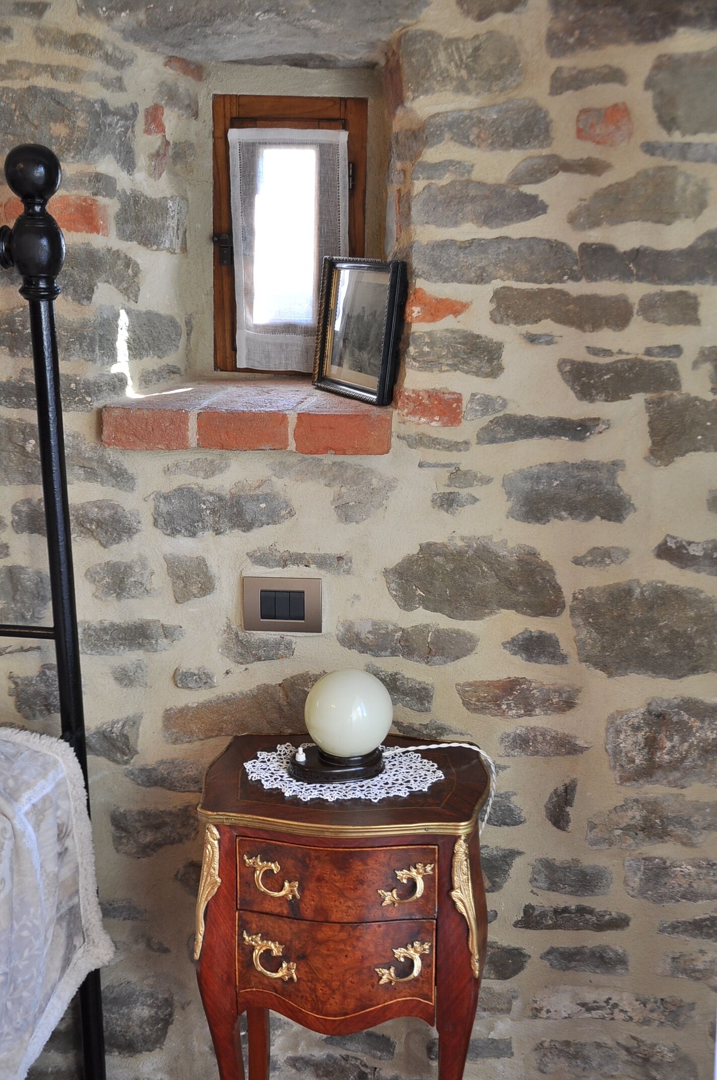 Una habitación con pared de piedra, puerta de madera, pequeña ventana, mesita de noche con un florero blanco y cama con cabecera negra.