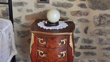 Una habitación con pared de piedra, puerta de madera, pequeña ventana, mesita de noche con un florero blanco y cama con cabecera negra.