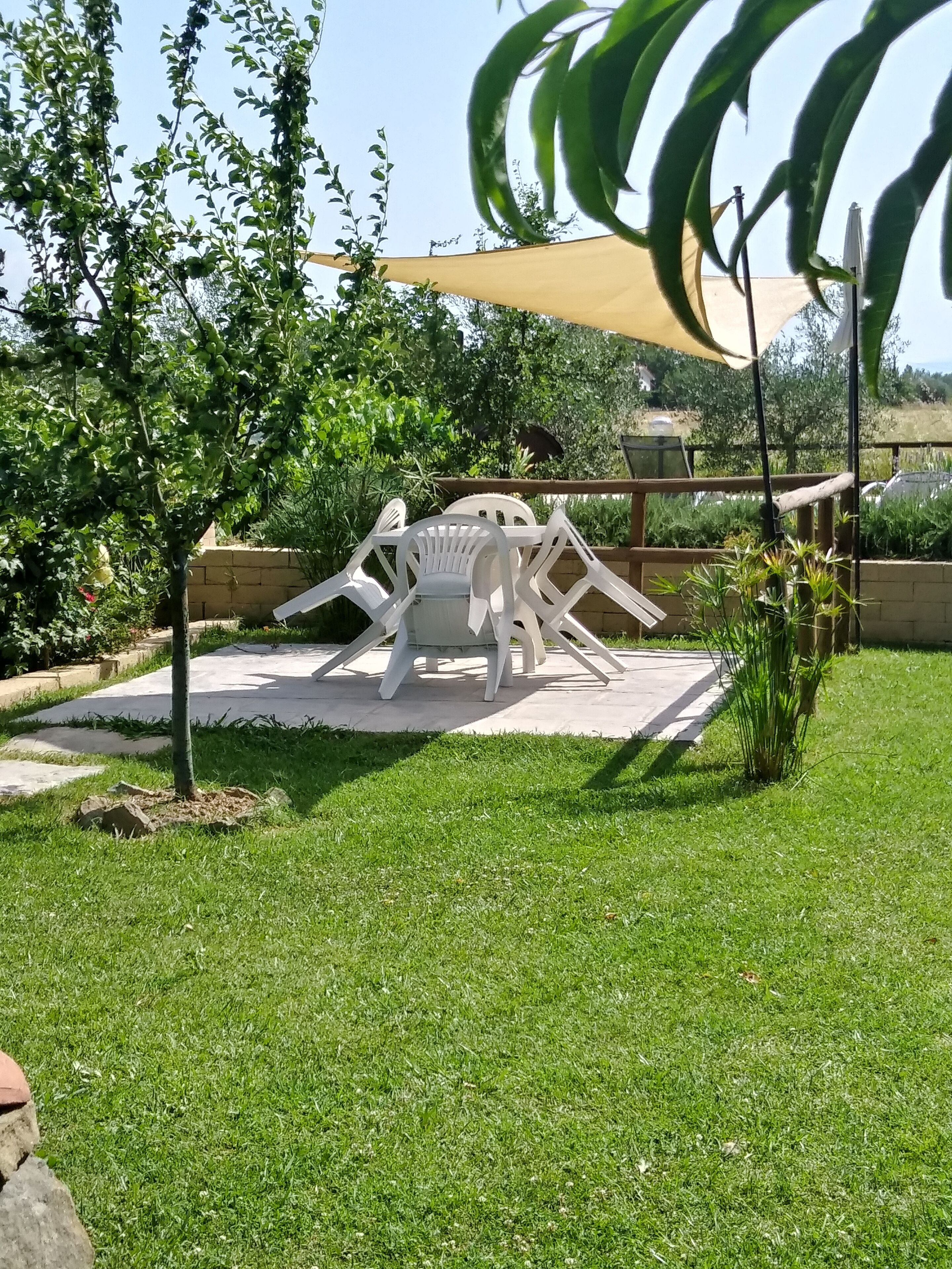 A patio area with a white plastic table and chairs, surrounded by greenery and a yellow shade sail.