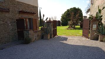 Un camino de grava que conduce a un edificio de piedra con una puerta metálica, un pequeño jardín con un árbol y una planta de cactus.