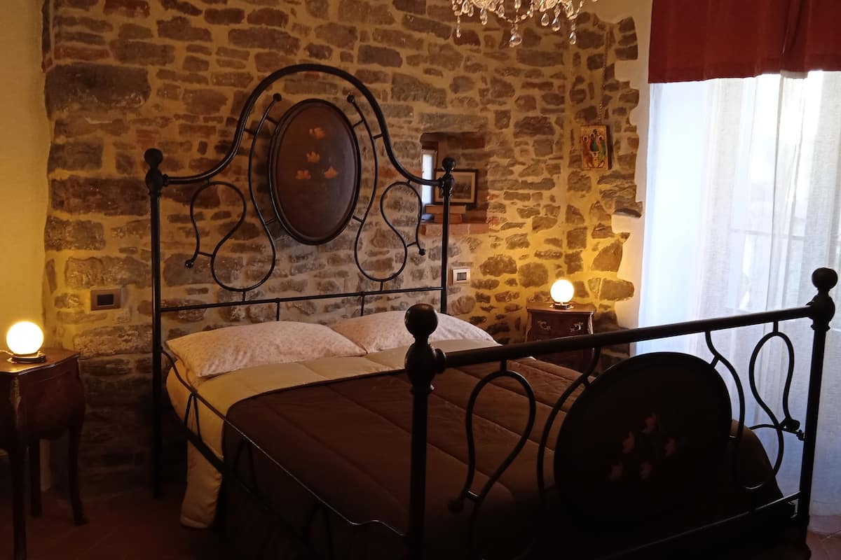 Un dormitorio con pared de piedra, una cama con cabecera, dos mesitas de noche con lámparas y una ventana con cortinas.