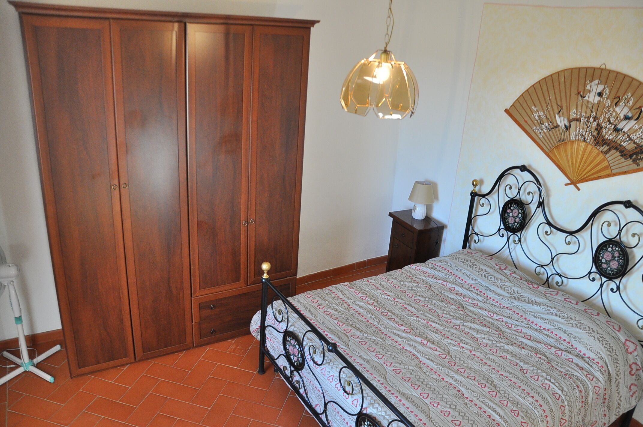 A bedroom with a wooden wardrobe, a bed with a patterned bedspread, a bedside table with a lamp, and a wall-mounted fan.