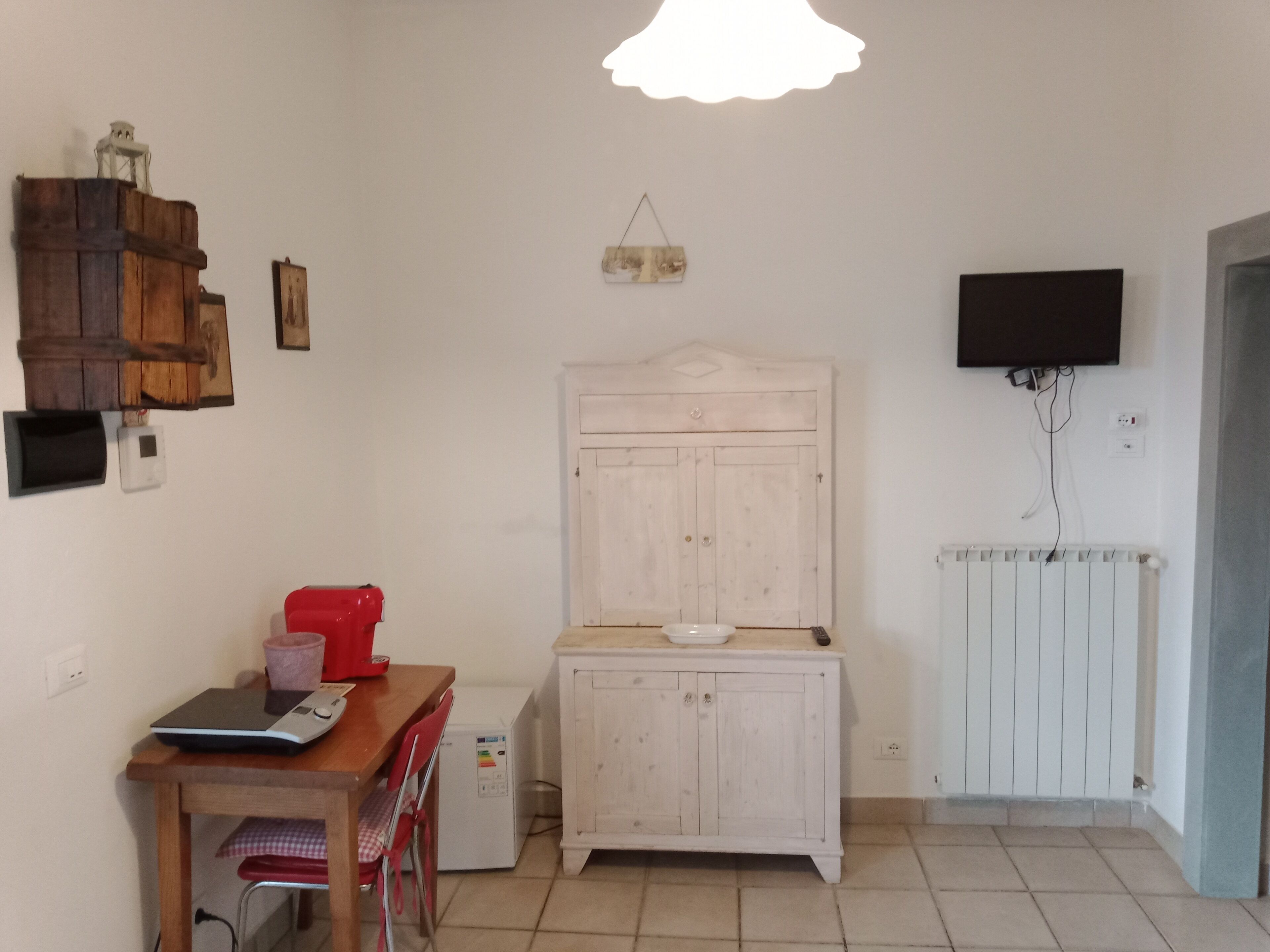 Une petite cuisine avec une table en bois, un réfrigérateur, un meuble blanc et un téléviseur fixé au mur.
