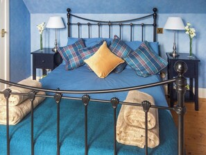 A bed with a metal frame, blue bedding, and plaid pillows. There are two bedside tables with lamps and vases of flowers.