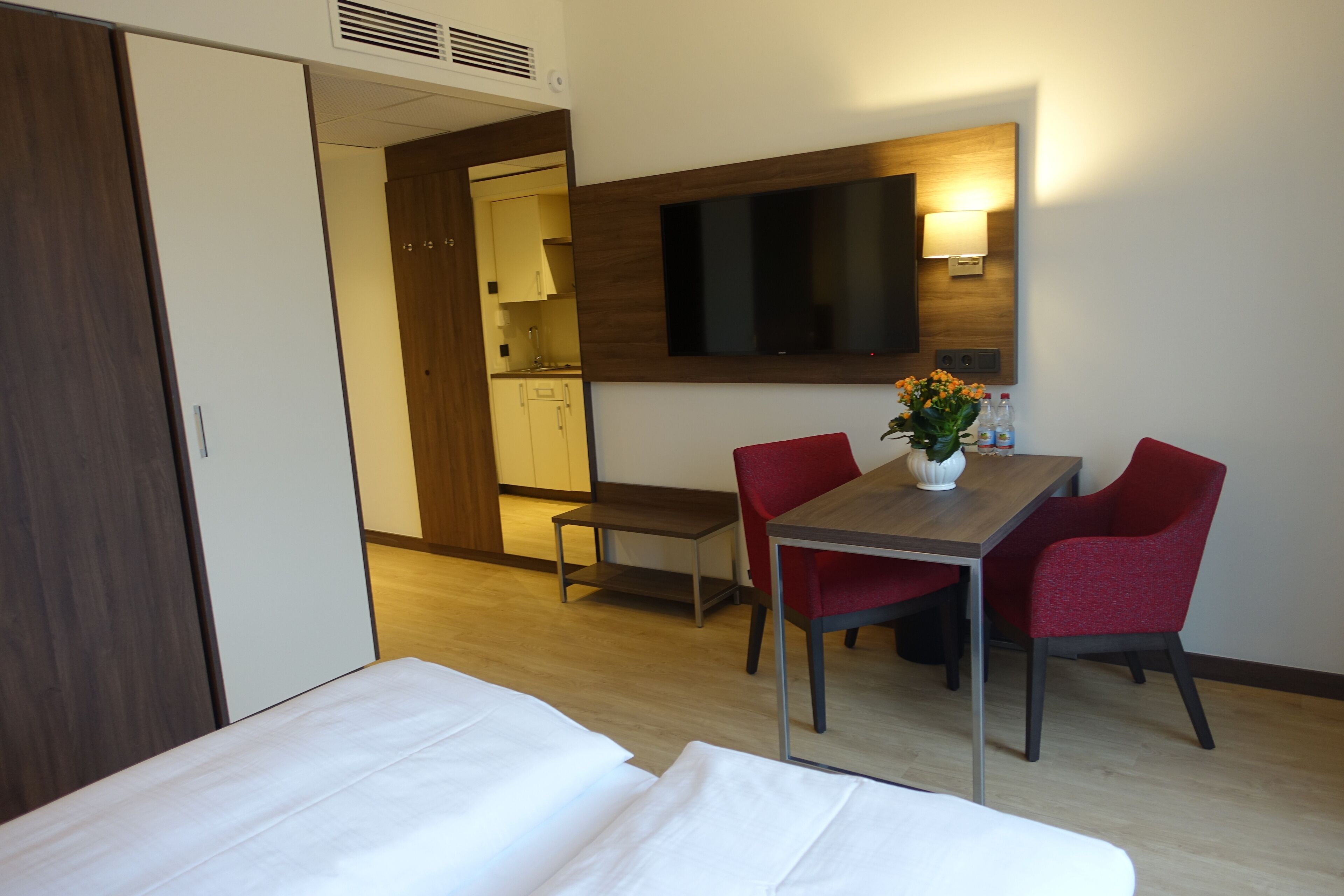 Una habitación de hotel con cama, escritorio, dos sillas rojas, televisor montado en la pared y una mesita con un jarrón de flores.