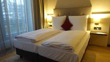 A double bed with white linens, two bedside tables with lamps, a red accent pillow, and a view of the outdoors through sheer curtains.