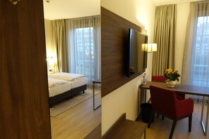 Quarto de hotel com cama, escrivaninha com cadeira, televisão e janela com cortinas.