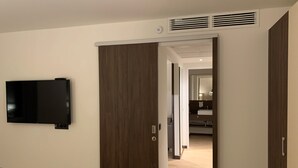 A modern hotel room with a flat-screen TV mounted on the wall, a wooden door, and a hallway leading to a bathroom with a mirror and sink. - Heidelberg Hospitality Boarding House (Heidelberg)