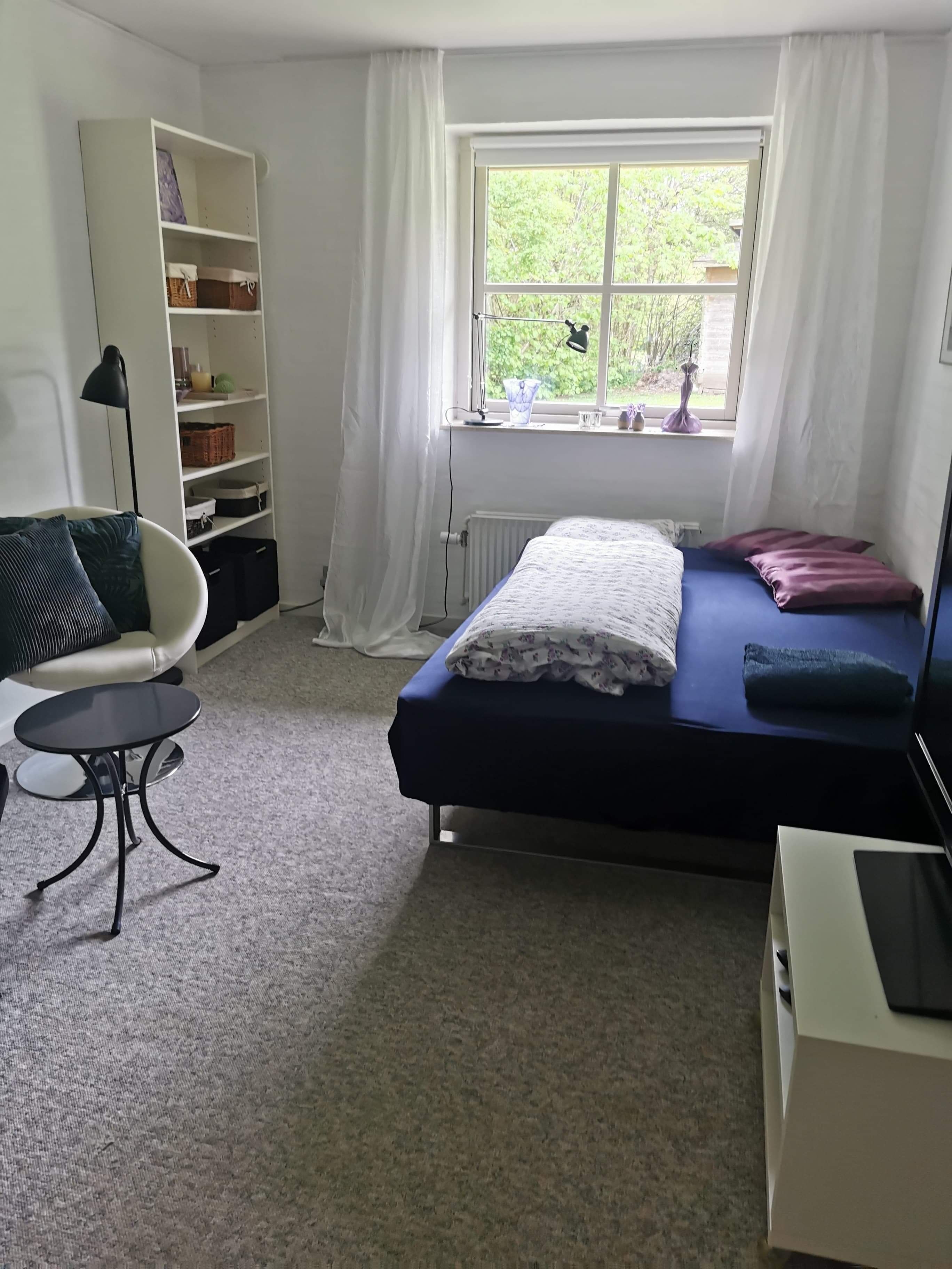 Un dormitorio con cama, una silla, un escritorio, una estantería y una ventana con vistas a zonas verdes.
