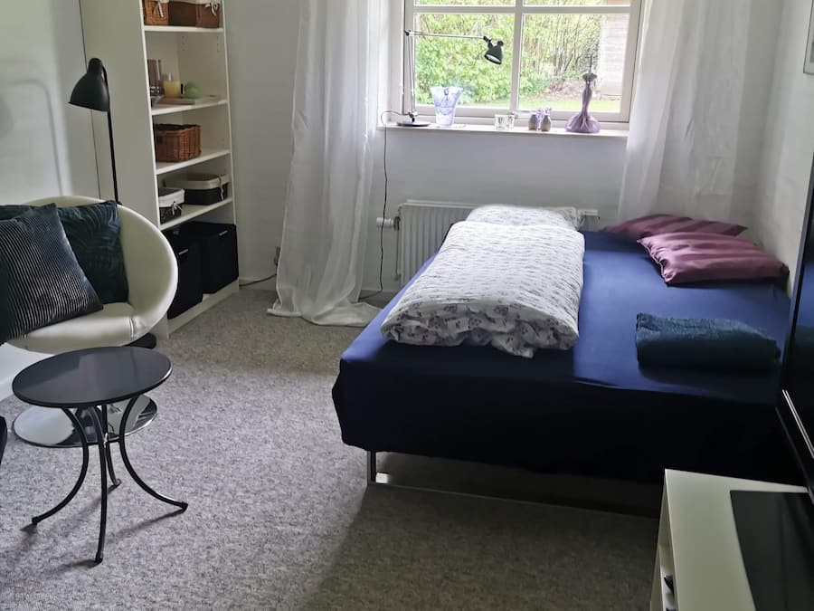 Un dormitorio con cama, una silla, un escritorio, una estantería y una ventana con vistas a zonas verdes.