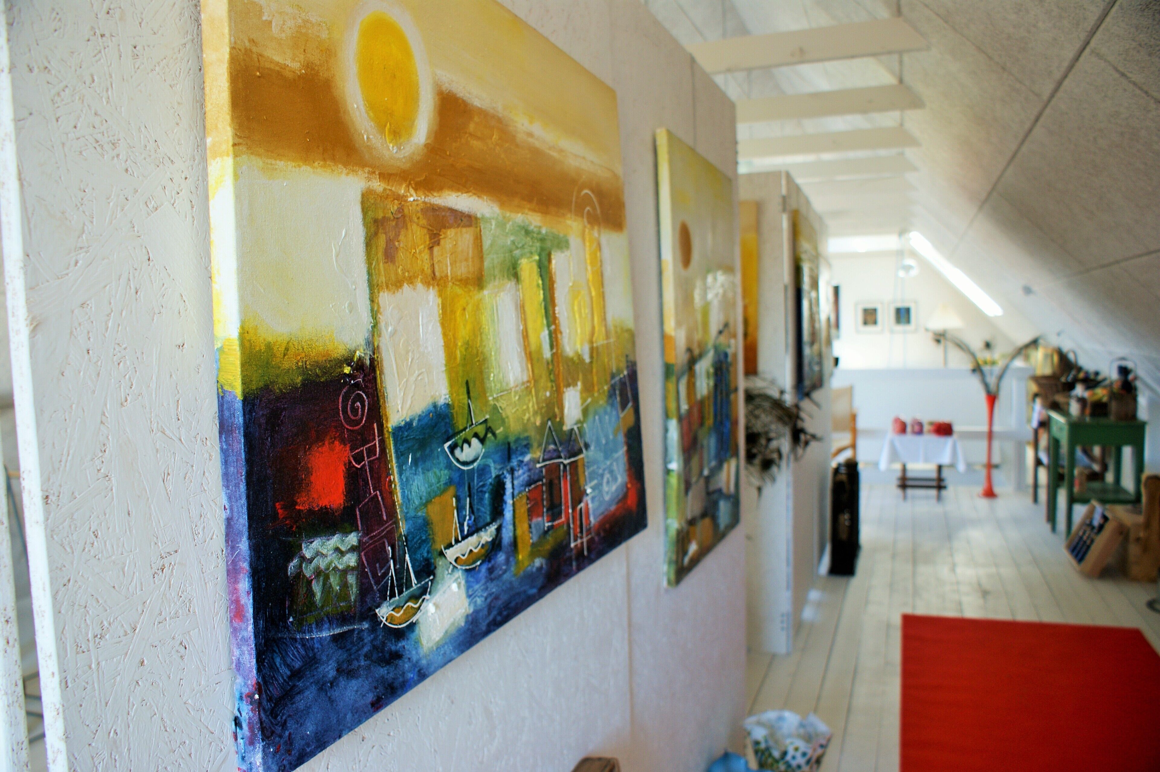 Un couloir aux murs ornés de peintures abstraites aux couleurs vives, un tapis rouge et, au fond, une table recouverte d’une nappe blanche.