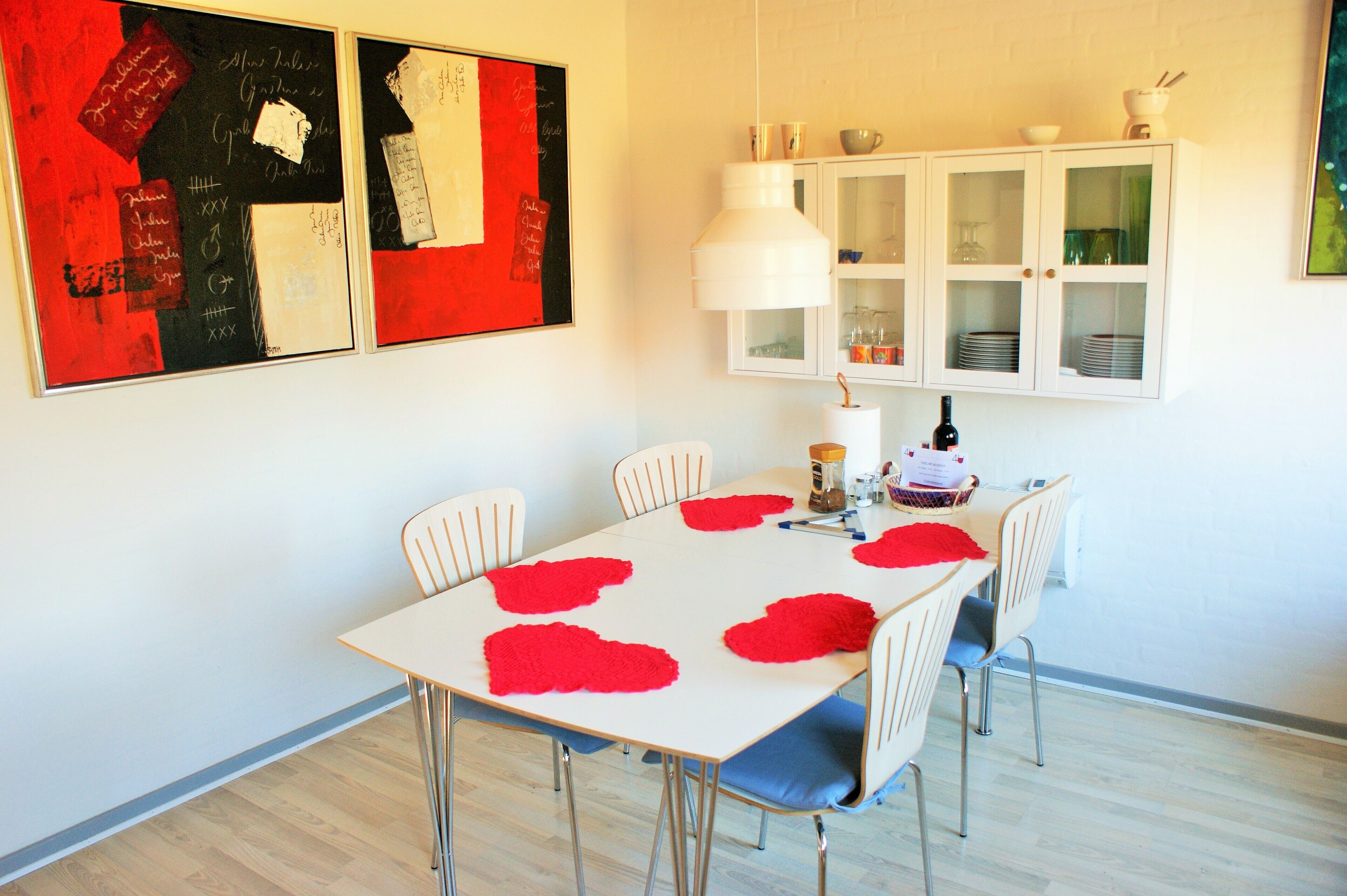 A dining area with a white table, red heart-shaped placemats, and a white cabinet with glass doors.