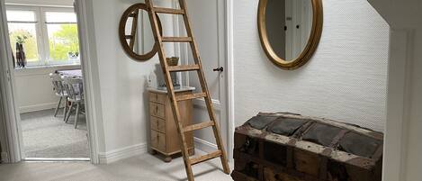 A room with a wooden ladder leaning against a door, a round mirror on the wall, and a rustic chest of drawers.