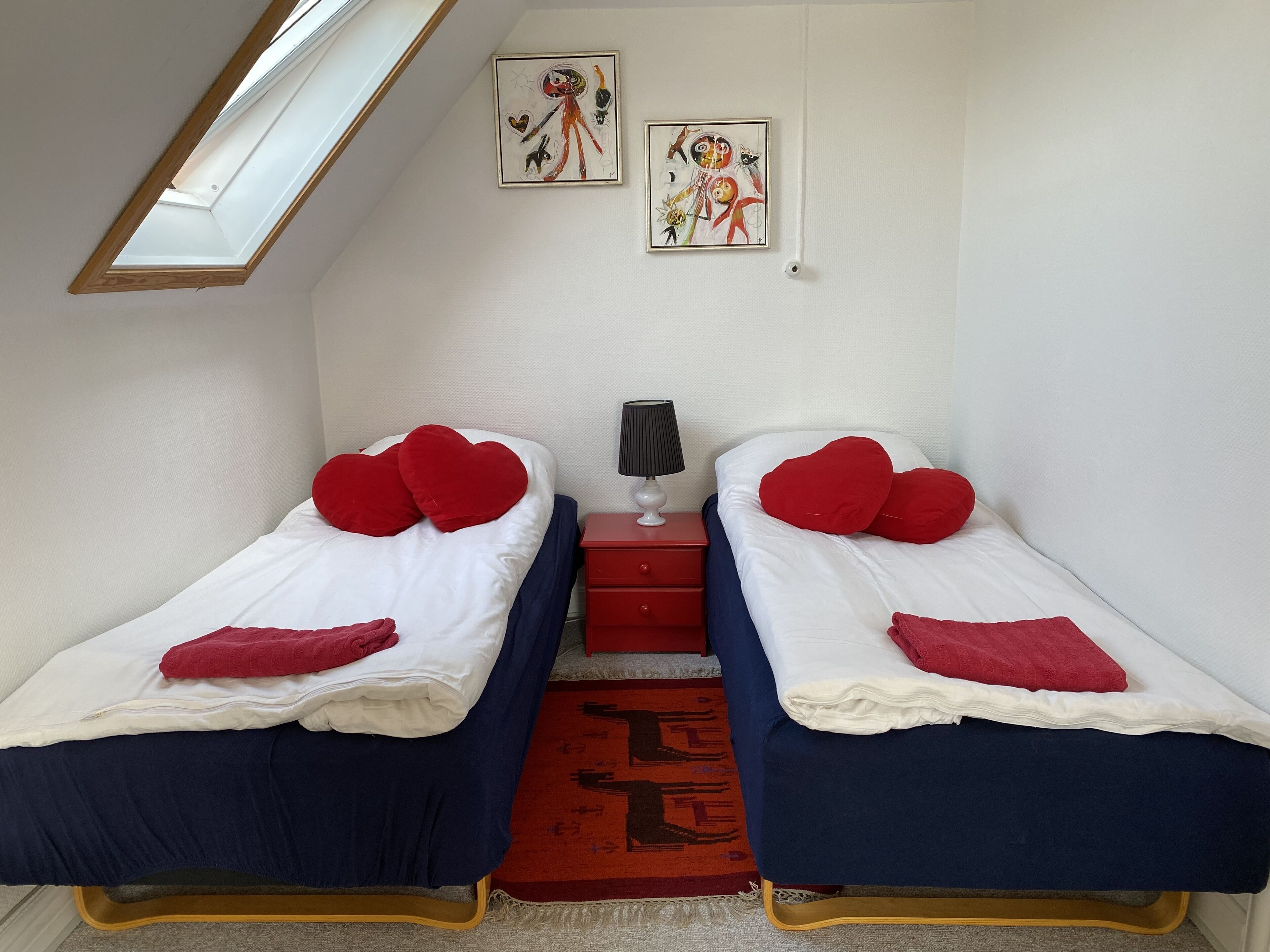 A bedroom with two single beds, each with red heart-shaped pillows and a blue blanket. There is a small bedside table with a lamp, and a red rug on the floor.