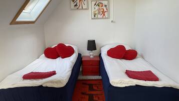 A bedroom with two single beds, each with red heart-shaped pillows and a blue blanket. There is a small bedside table with a lamp, and a red rug on the floor.