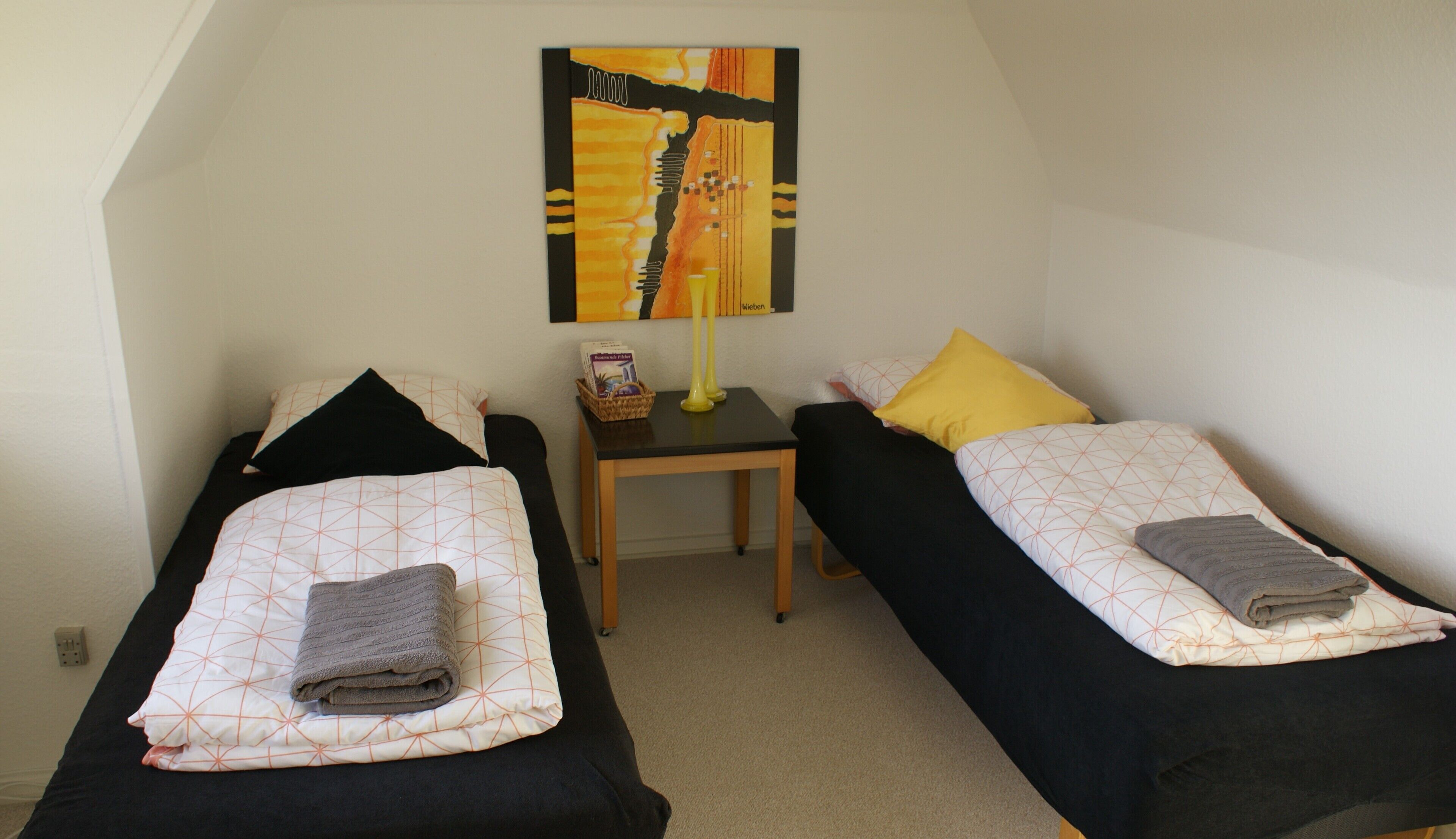 A room with two single beds, a small table, a lamp, and a painting on the wall.