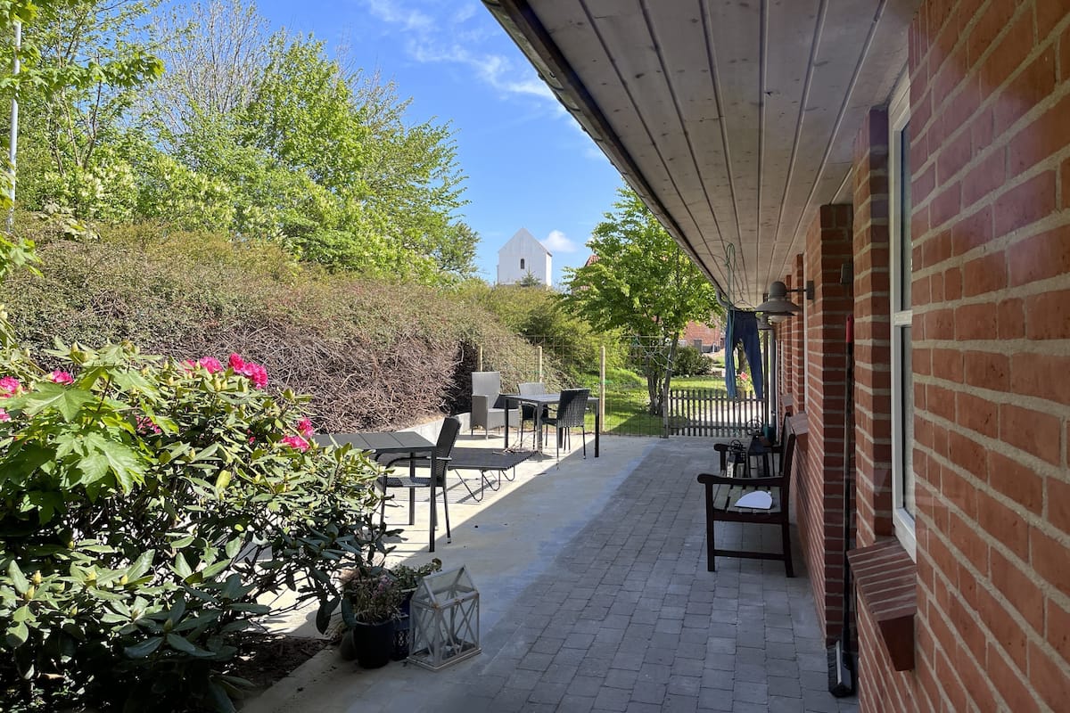 Eine überdachte Terrasse mit Sitzgelegenheiten im Freien, einer Ziegelwand und Blick auf ein weißes Gebäude in der Ferne.
