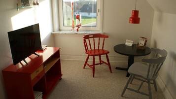 Una habitación con televisión, una silla roja, una silla gris, una mesita y una ventana con vista a una casa y árboles.