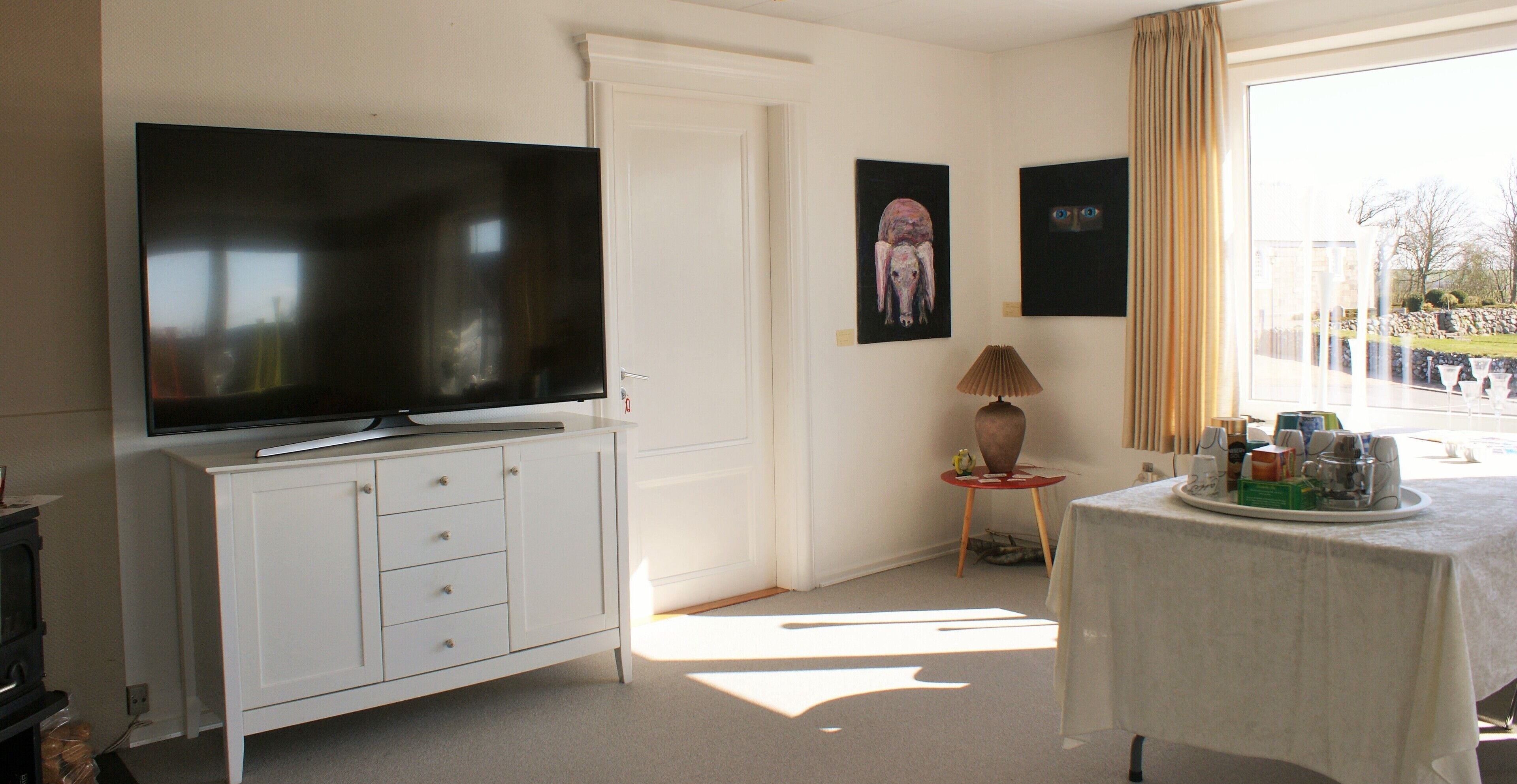 Quarto com televisão de tela plana em um armário branco, uma mesa pequena com abajur e obras de arte na parede.