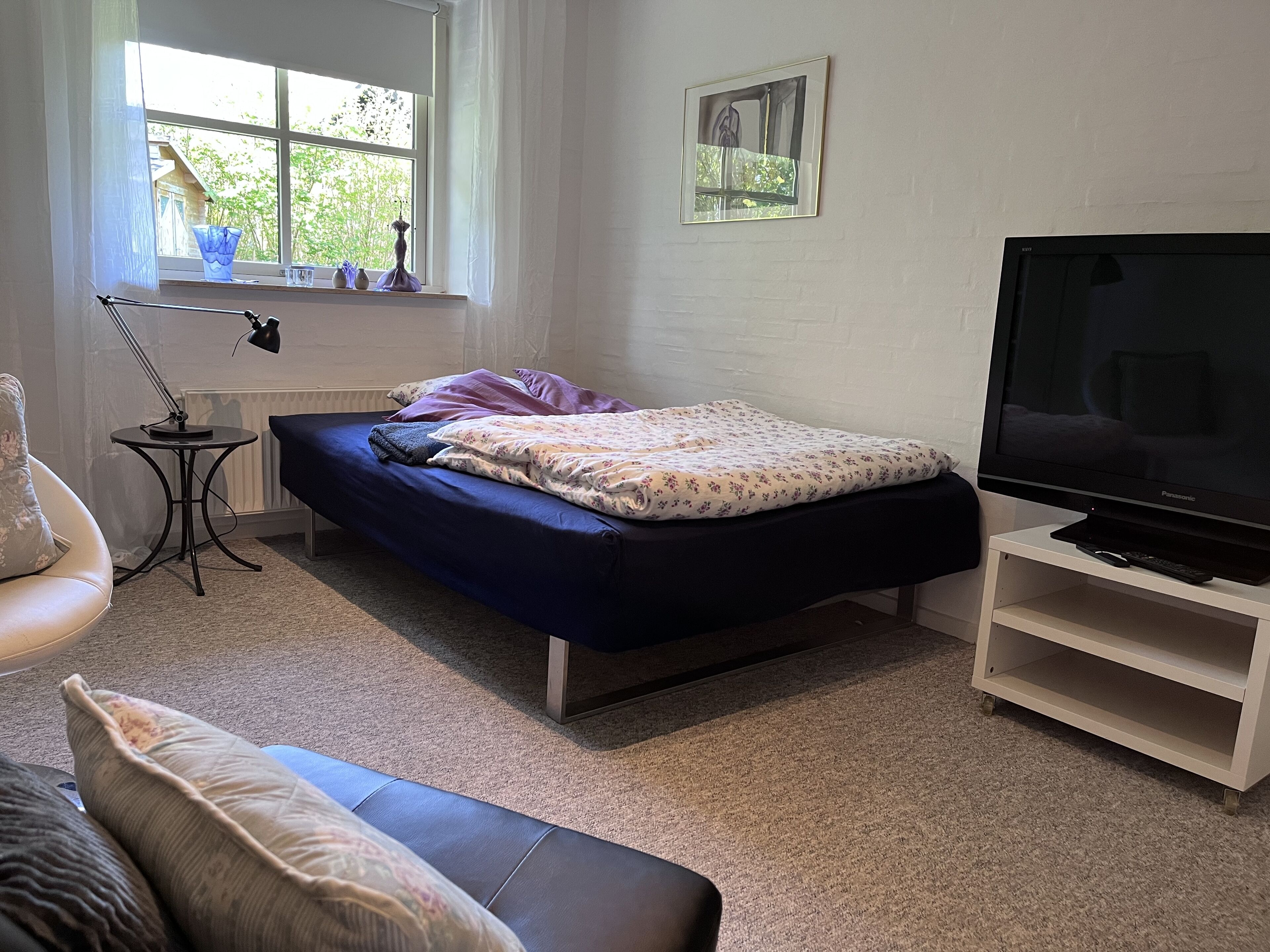 Ein Schlafzimmer mit einem Bett, einem Fernseher auf einem Schrank, einem Fenster mit Vorhängen und einem kleinen Tisch mit einer Lampe.