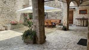 Un patio de piedra con una mesa y sillas bajo un arco, rodeado de muros de piedra y macetas con plantas.