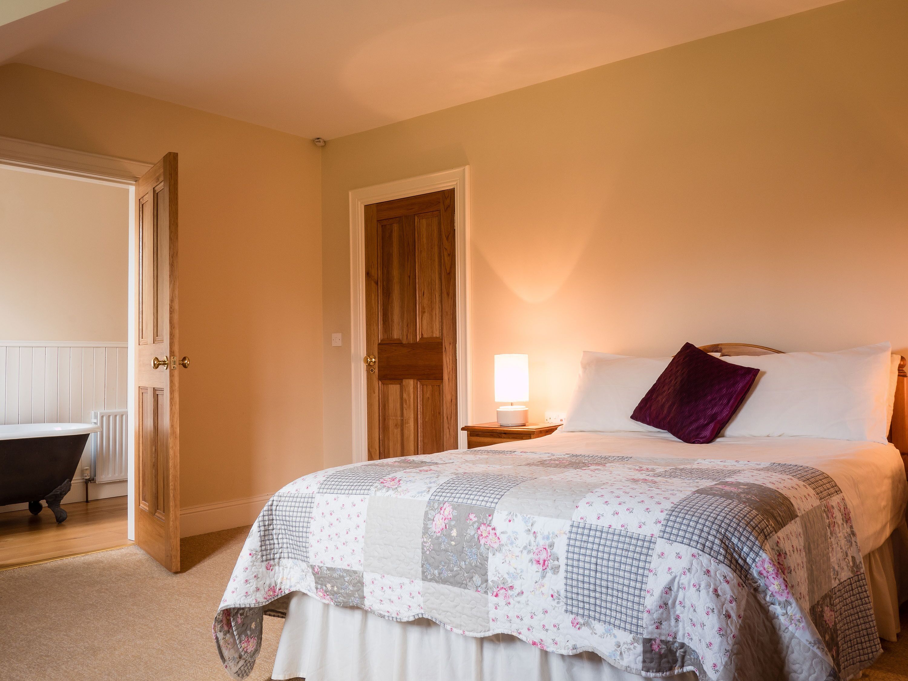 Ein Schlafzimmer mit Bett, Nachttisch, Lampe, Holztür und einem Badezimmer mit Badewanne.