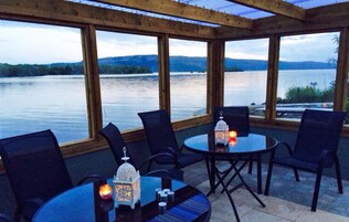 A covered outdoor seating area with tables and chairs, overlooking a lake with distant hills.