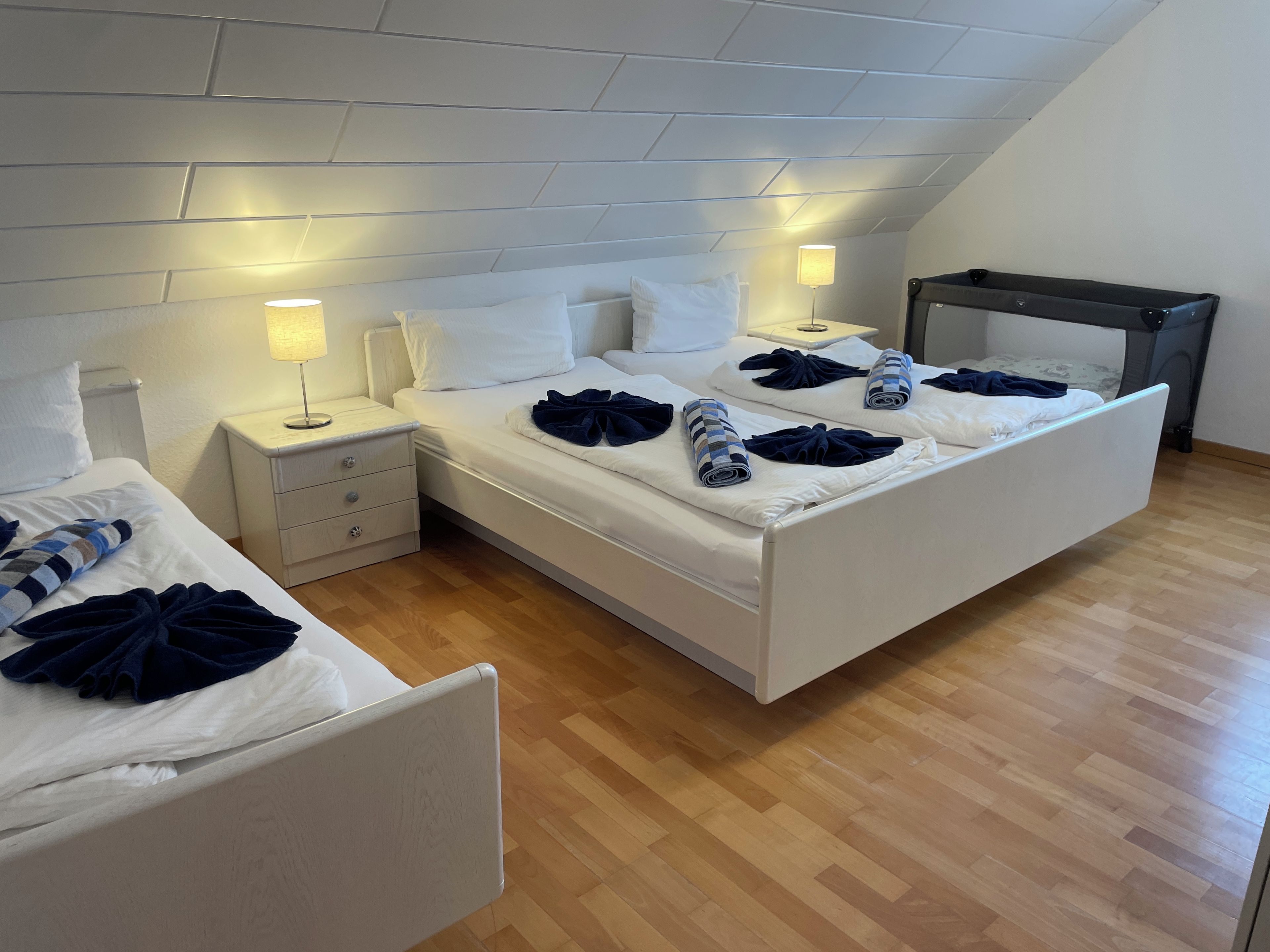 A modern hotel room with a large bed, a smaller bed, a nightstand with a lamp, and a ceiling with a sloped design.
