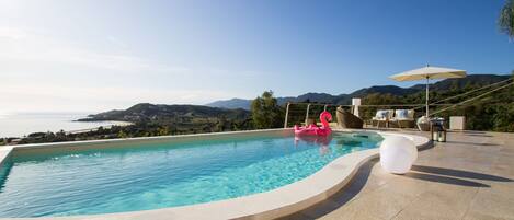 Una piscina con vistas al mar y a las montañas, un flotador de flamenco rosa y mobiliario exterior con sombrillas.
