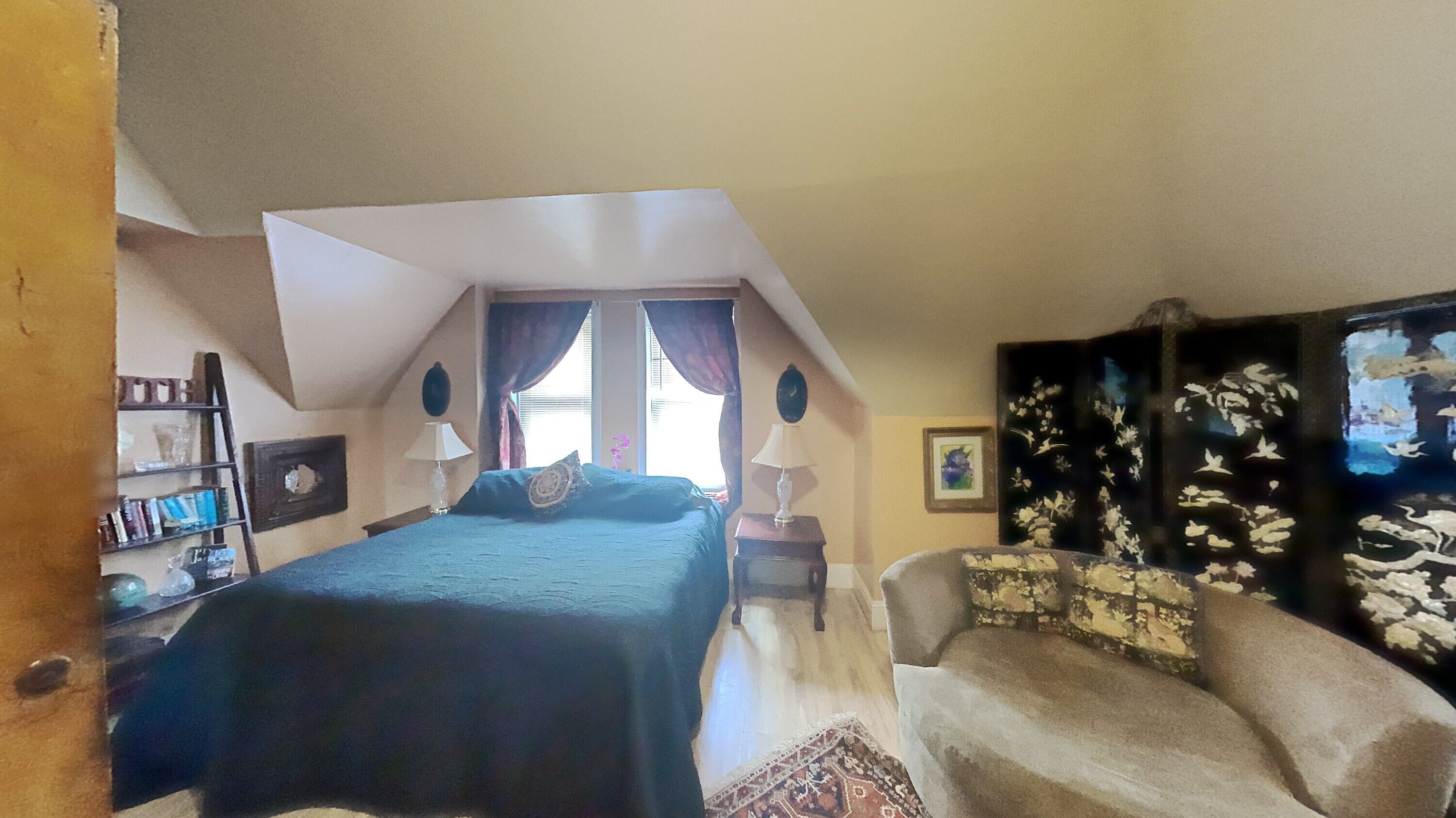 A bedroom with a bed, a chair, a nightstand, a window with curtains, a bookshelf, and a wall with a painting.