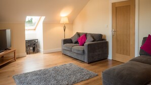 A cozy living room with a grey sofa, a wooden door, a TV on a stand, and a lamp. - The Apartment at Mullans Bay (Enniskillen)