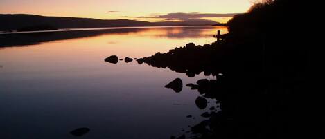 A calm lake at sunset with a rocky shoreline and distant hills.