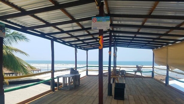 Un área de playa techada con una terraza de madera, mesas, sillas y vistas al océano.