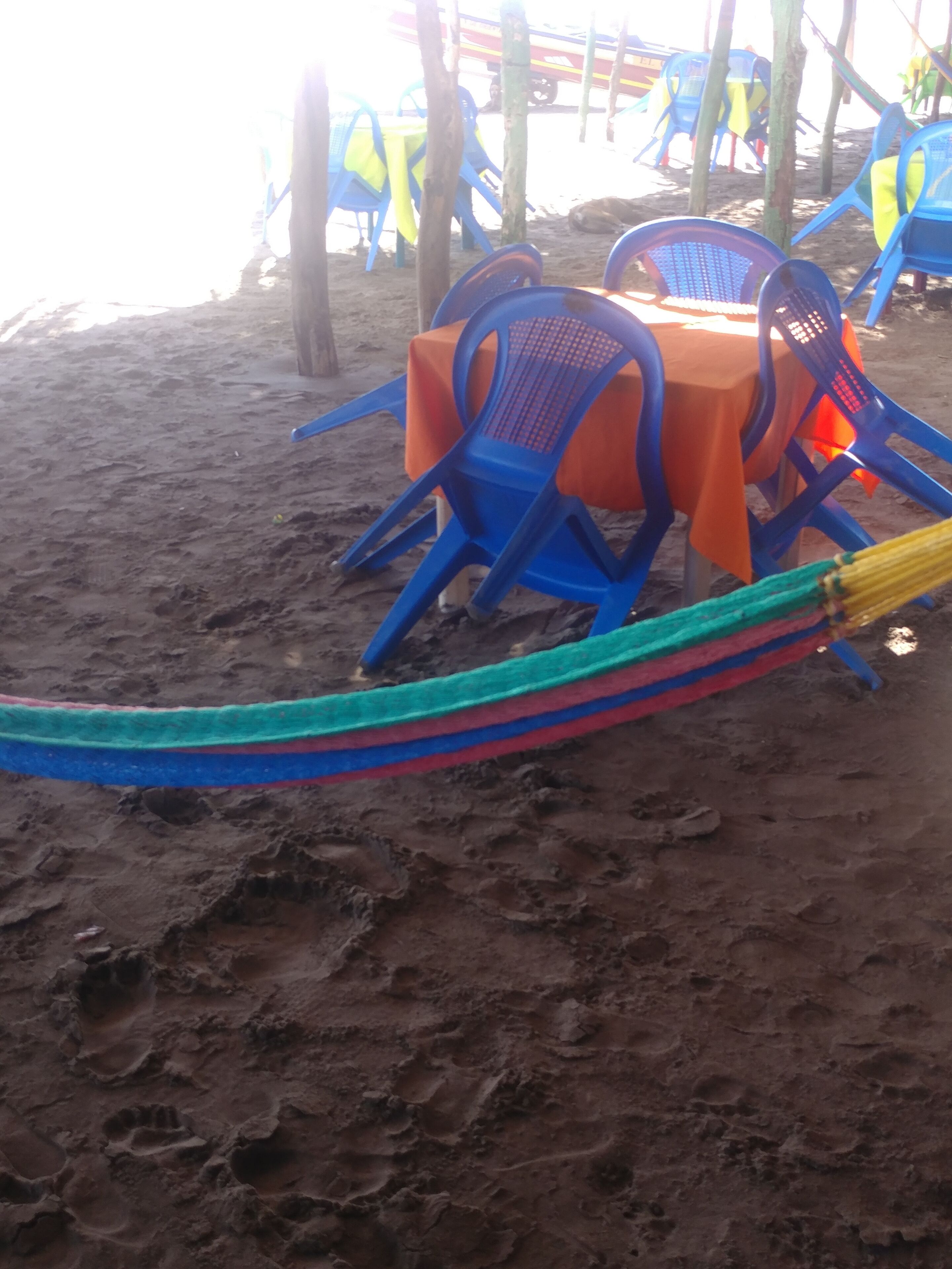 A beach setting with blue and orange plastic chairs and tables, a hammock, and a sandy ground.